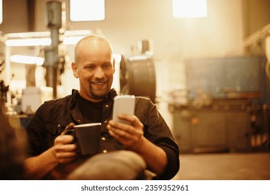 Middle aged car mechanic using a smartphone and drinking coffee while on a break in the mechanics shop - Powered by Shutterstock