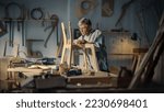Middle Aged Carpenter Putting on Glasses, Reading Blueprints and Starting to Assemble Parts of a Wooden Chair. Stylish Furniture Designer Working in a Studio in Loft Space with Tools on the Walls.