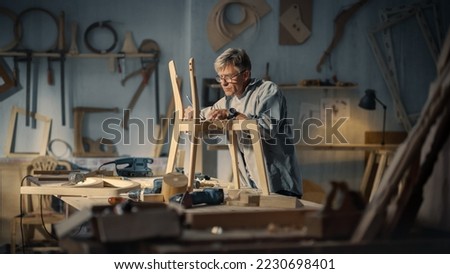 Middle Aged Carpenter Putting on Glasses, Reading Blueprints and Starting to Assemble Parts of a Wooden Chair. Stylish Furniture Designer Working in a Studio in Loft Space with Tools on the Walls.
