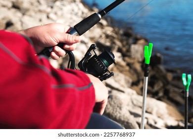 Middle aged caucasian male in red outfit is sitting on the river coast with fishing rod, holding it in left hand. Sunny morning in summer. Close up view of hands and reel of rod - Shutterstock ID 2257768937