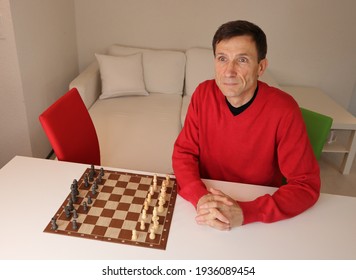 A Middle Aged Caucasian Man Sitting Alone At A Table With A Chess Set And An Empty Red Chair Next To Him. He Has A Loney Expression.