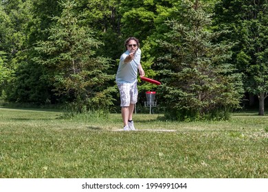 Middle aged Caucasian man in tee shirt, shorts, and sunglasses playing disc golf. Head on view of him tossing red disc with disc golf basket in the background.
