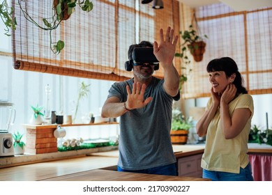 A Middle Aged Caucasian Man Is Using A Virtual Reality Headset With His Wife Next To Him Laughing At Him Being Silly