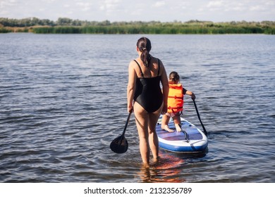 Middle aged Caucasian mother looking after little daughter sup boarding on lake with oar in hands in life jacket. Active holidays full of adventures. Inculcation of love for sports from childhood. - Shutterstock ID 2132356789