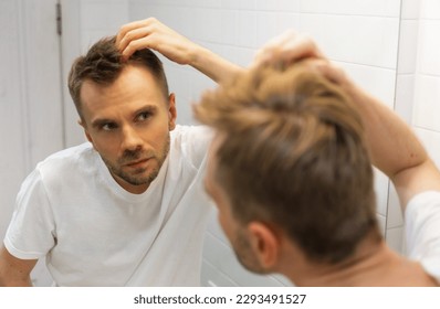 Middle aged caucasian white man with a short beard looks at his hair in the mirror in the bathroom and worried about balding. The concept of the problem of male hair loss, early baldness and alopecia.