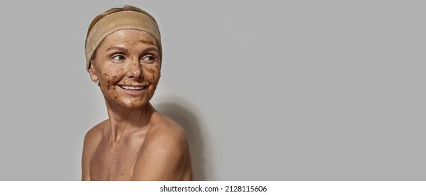 Middle aged caucasian woman with coffee and salt scrub on her face looking away. Smiling blonde female wearing headband. Body and skin care. Cosmetology and spa. White background in studio. Copy space