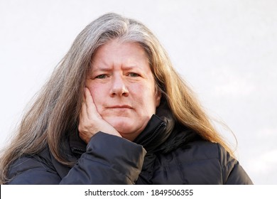 Middle Aged Caucasian Woman With Long Thick Hair In Puffy Jacket And Cold Weather Leans Chin On Hand With Furrowed Brow In Disapproval Outside                                