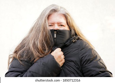 Middle Aged Caucasian Woman With Long Thick Hair In Puffy Jacket And Cold Weather Has Collar Zipped Up Over Face While Grabbing Jacket To Keep Warm                              