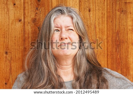 Middle aged caucasian woman with long gray and brown hair against a wooden wall looks up and to the right with a goofy expression                              