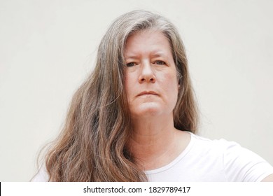 Middle aged caucasian woman with thick, long brown hair, wearing a white shirt against a white background, looks stern while looking at the camera                               
