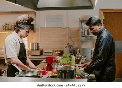 Middle aged chef dressed in black uniform and his young sous chef cooking dishes together in modern restaurant kitchen - Powered by Shutterstock