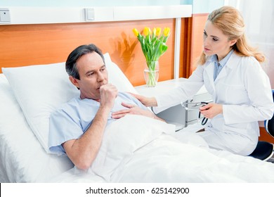 Middle Aged With Chest Pain Lying On Bed And Doctor With Stethoscope Sitting Near Him In Hospital 