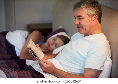 Middle Aged Couple In Bed Together With Man Reading Book