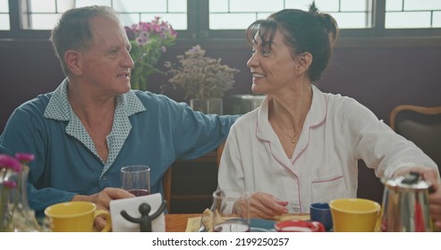 Middle Aged Couple Having Breakfast Together. Older Couple Wearing Pajamas Talking And Smiling. Senior Woman Drinking Coffee. Husband Drinking Orange Juice.