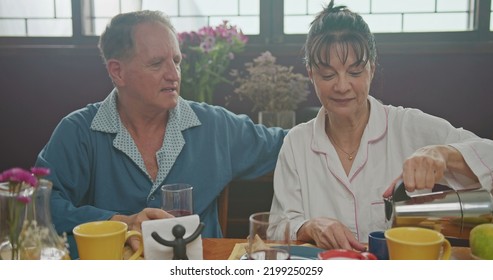 Middle Aged Couple Having Breakfast Together. Older Couple Wearing Pajamas Talking And Smiling. Senior Woman Drinking Coffee. Husband Drinking Orange Juice.