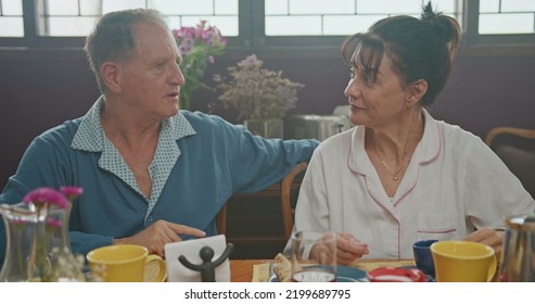 Middle Aged Couple Having Breakfast Together. Older Couple Wearing Pajamas Talking And Smiling. Senior Woman Drinking Coffee. Husband Drinking Orange Juice.