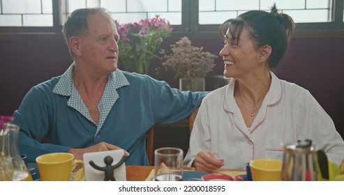 Middle Aged Couple Having Breakfast Together. Older Couple Wearing Pajamas Talking And Smiling. Senior Woman Drinking Coffee. Husband Drinking Orange Juice.