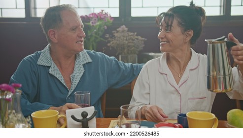Middle Aged Couple Having Breakfast Together. Older Couple Wearing Pajamas Talking And Smiling. Senior Woman Drinking Coffee. Husband Drinking Orange Juice.