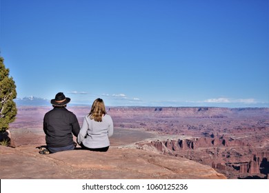 Middle Aged Couple Sitting On The Edge Of Canyonlands Canyon Looking Off Into The Distance.  