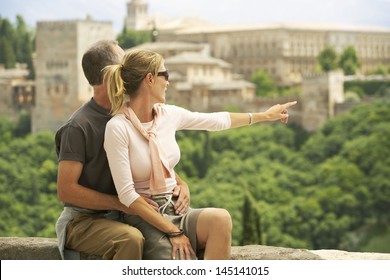 Middle aged couple sitting on wall with woman pointing at view; Granada Spain