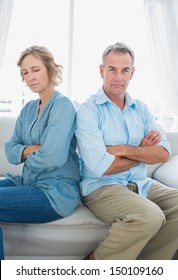 Middle Aged Couple Sitting On The Sofa Not Speaking After A Dispute With Man Looking At Camera At Home In The Living Room