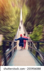 Middle Aged Couple Walking On Suspension Bridge With Their Back Turned Towards Camera. Radial Zoom Effect Defocusing Filter Applied, With Vintage Instagram Look.