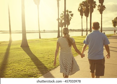 Middle Aged Couple Walking Together Holding Hands Along The Beach Boardwalk. Abstract Image Of Building Relationships And Marital Bonds. Early Evening Sunset And Warm Summer Tone