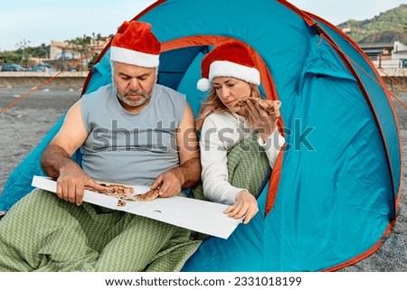 Middle aged couple wearing santa hats having christmas picnic on winter beach. Bearded man and pretty blond woman sitting in tent, talking and eating pizza.