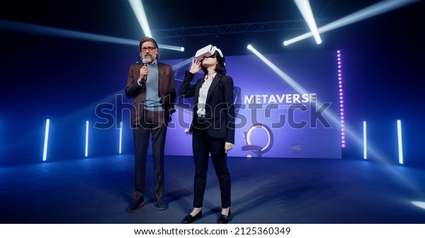 A\
middle aged creator demonstrating new augmented reality glasses\
technology to a female host and audience on stage, during a meta\
universe event in a contemporary studio with LED\
screen