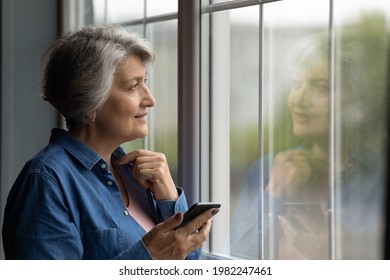 Middle aged dreamy woman holding cellphone standing near window looking outside. Grey haired older female pose indoor distracted from device usage staring outdoor. Older gen use modern tech concept - Powered by Shutterstock