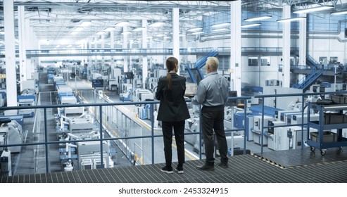 Middle Aged Engineer and Female Scientist Standing with Their Back, Using Laptop Computer and Talking in a Factory Facility with Equipment Producing Modern Technical Devices for Different Industries