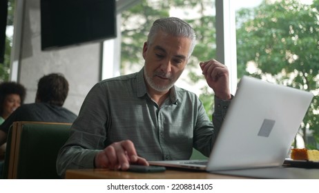 Middle Aged Entrepreneur Working At Coffee Shop In Front Of Laptop. Senior Male Executive Typing On Computer Keyboard Checking Phone And Using Modern Technology