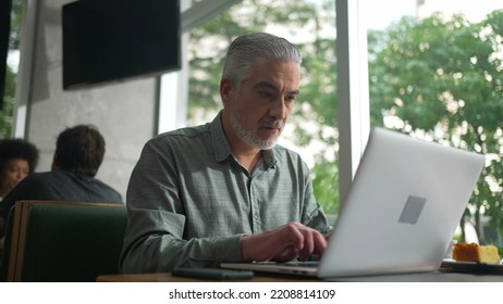 Middle Aged Entrepreneur Working At Coffee Shop In Front Of Laptop. Senior Male Executive Typing On Computer Keyboard Checking Phone And Using Modern Technology