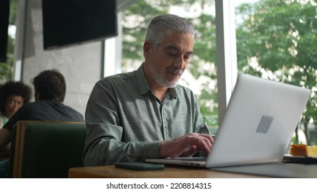 Middle Aged Entrepreneur Working At Coffee Shop In Front Of Laptop. Senior Male Executive Typing On Computer Keyboard Checking Phone And Using Modern Technology