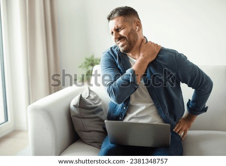 Middle Aged European Man Suffering From Neckache And Backache After Online Work On Laptop Computer, Sitting On Sofa At Home. Sick Guy With Earbuds Rubbing Body Inflamed Zones, Feeling Unwell