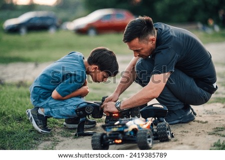 Middle aged father showing the issue with a wheel on his son's truck toy. They were playing with remote controlled toy cars and the boy's car overturned. The boy is carefully watching.