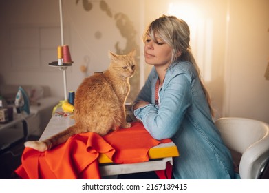 Middle Aged Female Fashion Designer Using Sewing Machine And Making A New Handmade Suit While Working At Home And Petting Her Pet Cat. Hobby Sewing As A Small Business Concept. Life With Pets.