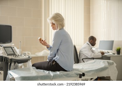 Middle Aged Female Patient Sitting On Couch And Looking At Bottle Of Prescription Drugs. Male General Practitioner Making Notes In Medical History In Background