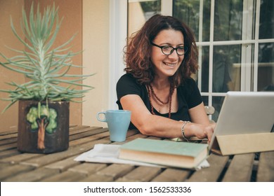 Middle aged female studying at home with books, newspaper and digital tablet pad. Woman reading a book and watching video online on new tech device. Education, modern lifestyle and leisure concept.