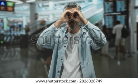 Middle aged grey haired man raises hands and smiles in busy airport concourse near escalator and flight screens, wearing glasses and light shirt; joyful travel optimism.