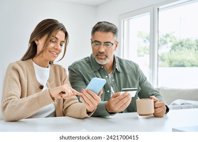 Middle aged happy couple older mature man and woman using smartphone looking at cell phone holding credit card buying online, making banking payment on ecommerce mobile shopping sitting at home table. - Powered by Shutterstock