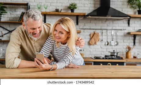 Middle Aged Happy Couple Using Mobile Phone While Standing In Kitchen At Home
