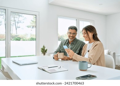 Middle aged happy couple using smartphone relaxing at living room table at home. Smiling mature older man and woman holding cellphone browsing internet, texting message on mobile cell phone technology - Powered by Shutterstock