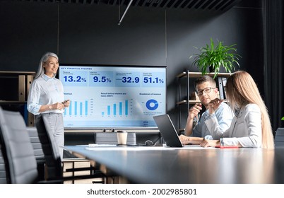 Middle Aged Happy Successful Asian Business Woman Ceo Executive Manager Presenting To Colleagues Trainees Interns Income Revenue Financial Data Research Results On Big Screen In Modern Office.