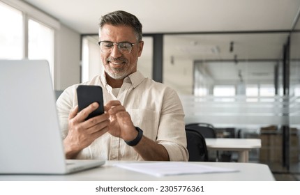 Middle aged Hispanic business manager ceo using cell phone mobile app, laptop. Smiling Latin or Indian mature man businessman holding smartphone sit in office working online on gadget with copy space. - Powered by Shutterstock