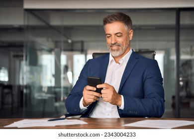 Middle aged Hispanic business manager ceo using cell phone mobile app, laptop. Smiling Latin or Indian mature man businessman holding smartphone sit in office working online on gadget with copy space. - Powered by Shutterstock