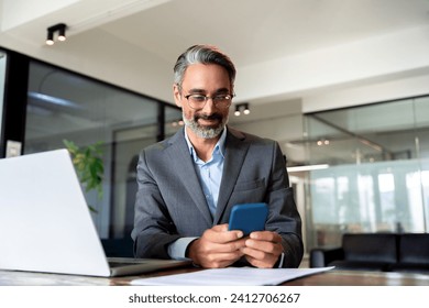 Middle aged Hispanic business manager ceo using cell phone mobile app, laptop. Smiling Latin or Indian mature man businessman holding smartphone sit in office working online on gadget with copy space. - Powered by Shutterstock