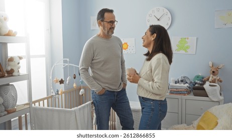 Middle aged hispanic couple standing in cozy bedroom smiling at each other near baby's cradle surrounded by soft toys and pastel decor, embodying love and family warmth.