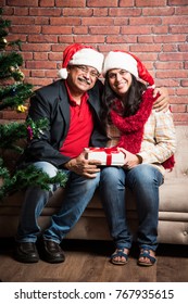 Middle Aged Indian Smart Couple Celebrating Christmas And Exchanging Gifts While Sitting On Sofa