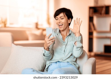 Middle aged lady using her mobile phone for online video chat, calling remote friend by smartphone, comfortably sitting on sofa in cozy living room. Modern digital communication
